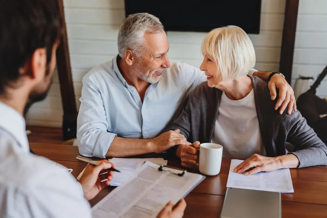 Smiling senior couple sitting together at a desk with paperwork and coffee, warmly interacting while meeting with a financial advisor who is partially visible in the foreground.