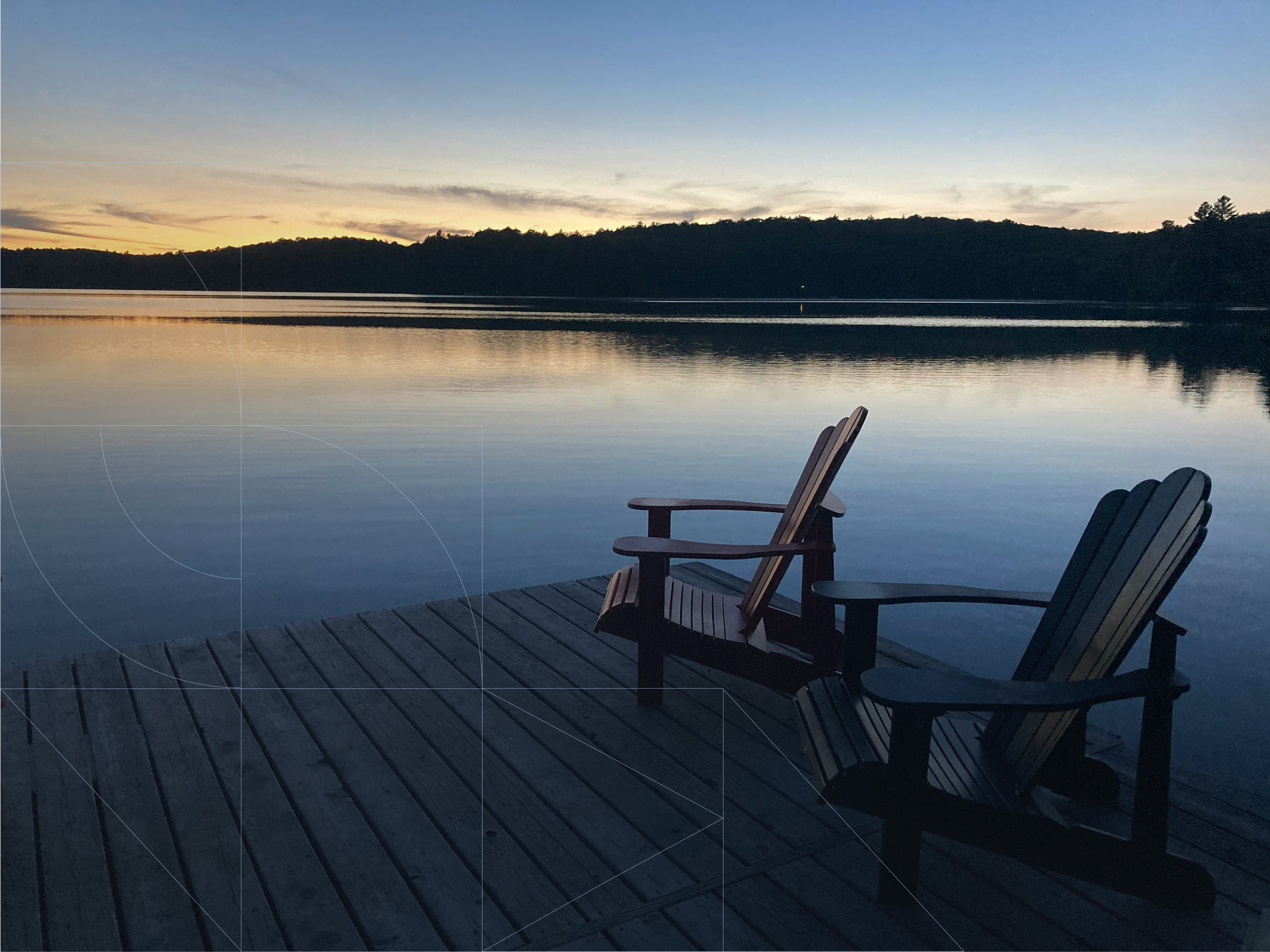 Calm lakeside scene at sunset with two wooden Adirondack chairs on a dock overlooking still water, reflecting the soft golden and blue evening sky and tree-lined horizon.