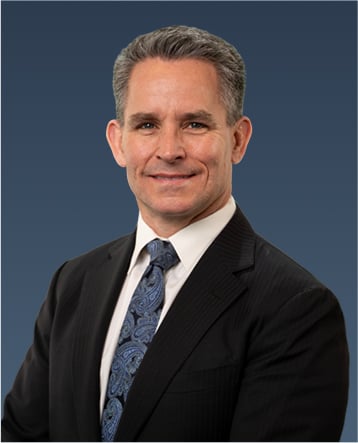 Professional portrait of a man with dark hair in a black suit and patterned blue tie, smiling slightly.