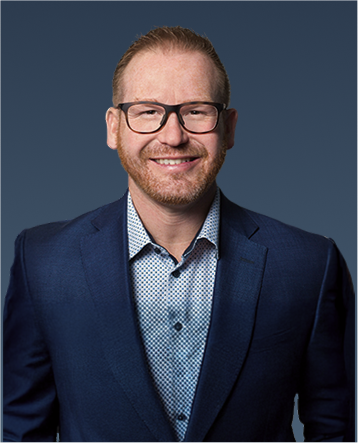 Professional portrait of a man with red hair, glasses, and a beard, wearing a blue blazer and light shirt, smiling.