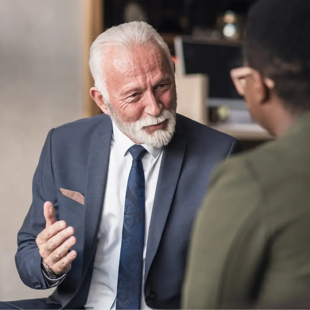 An older man with white hair and beard in a suit speaking to another person in a casual shirt.