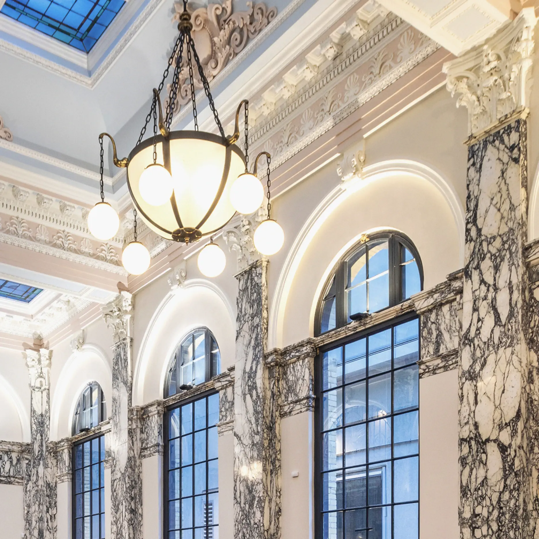 Interior of a grand building with tall arched windows, ornate marble columns, decorative molding, and a large chandelier with round lights hanging from the ceiling.