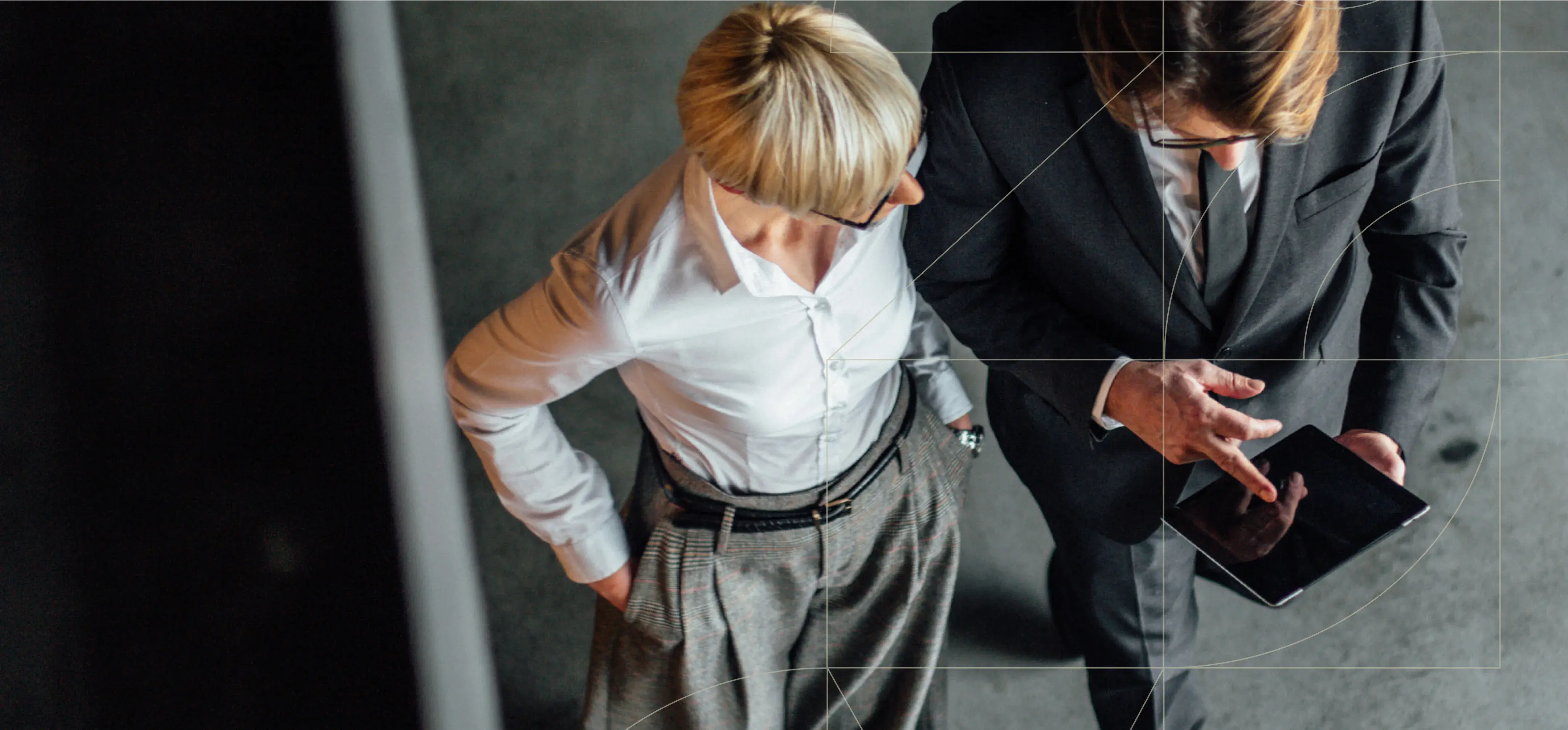 Two professionals stand together, looking at a tablet. One, in a white shirt and glasses, observes while the other, in a dark suit, points at the screen.