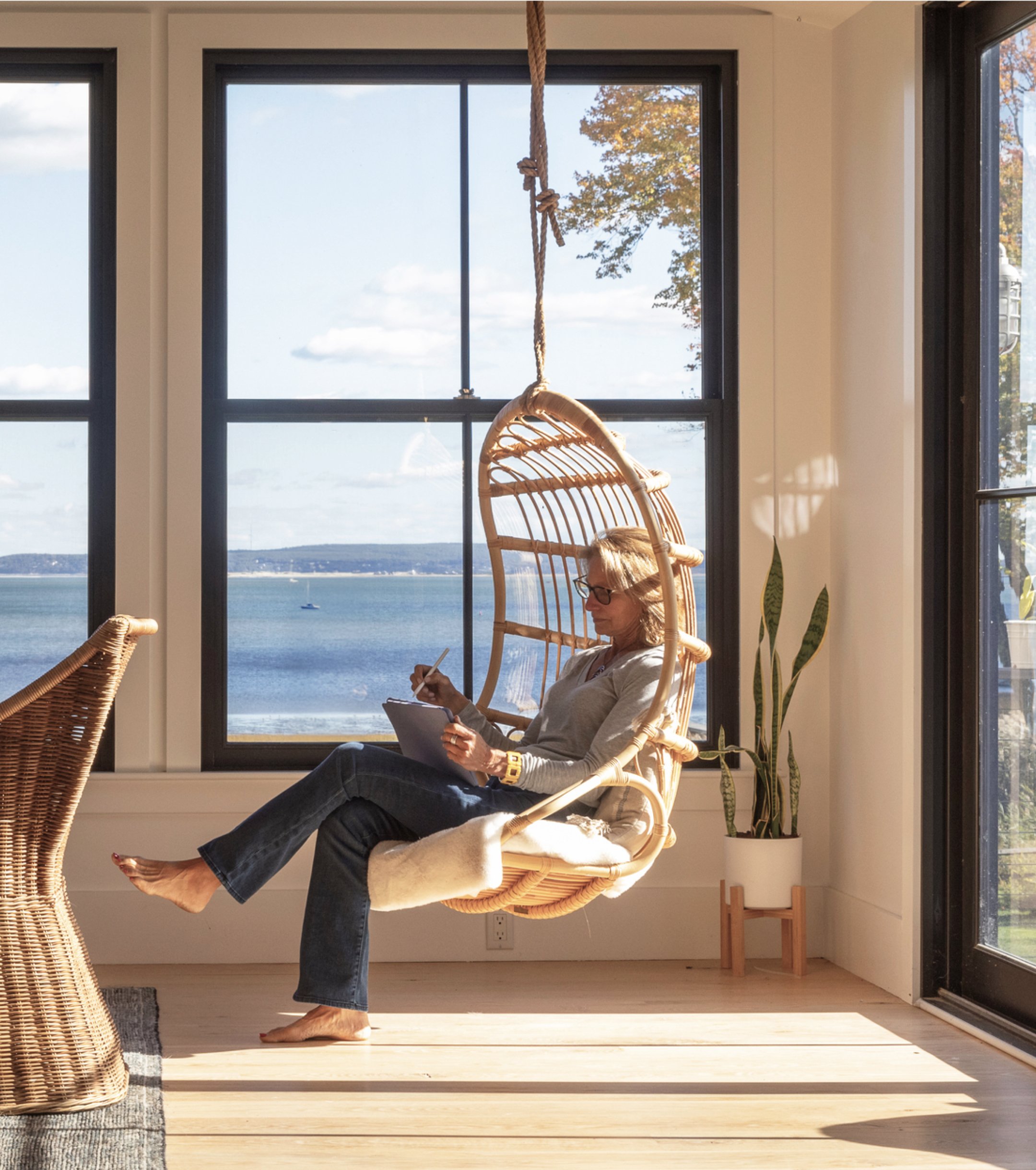 Person sits in a wicker hanging chair by a large window, writing in a notebook. Sunlight fills the room, with a view of the ocean and a flag outside.