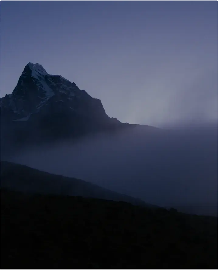 Dark mountain peak emerging above low-lying mist, with a soft purple-blue twilight sky in the background.