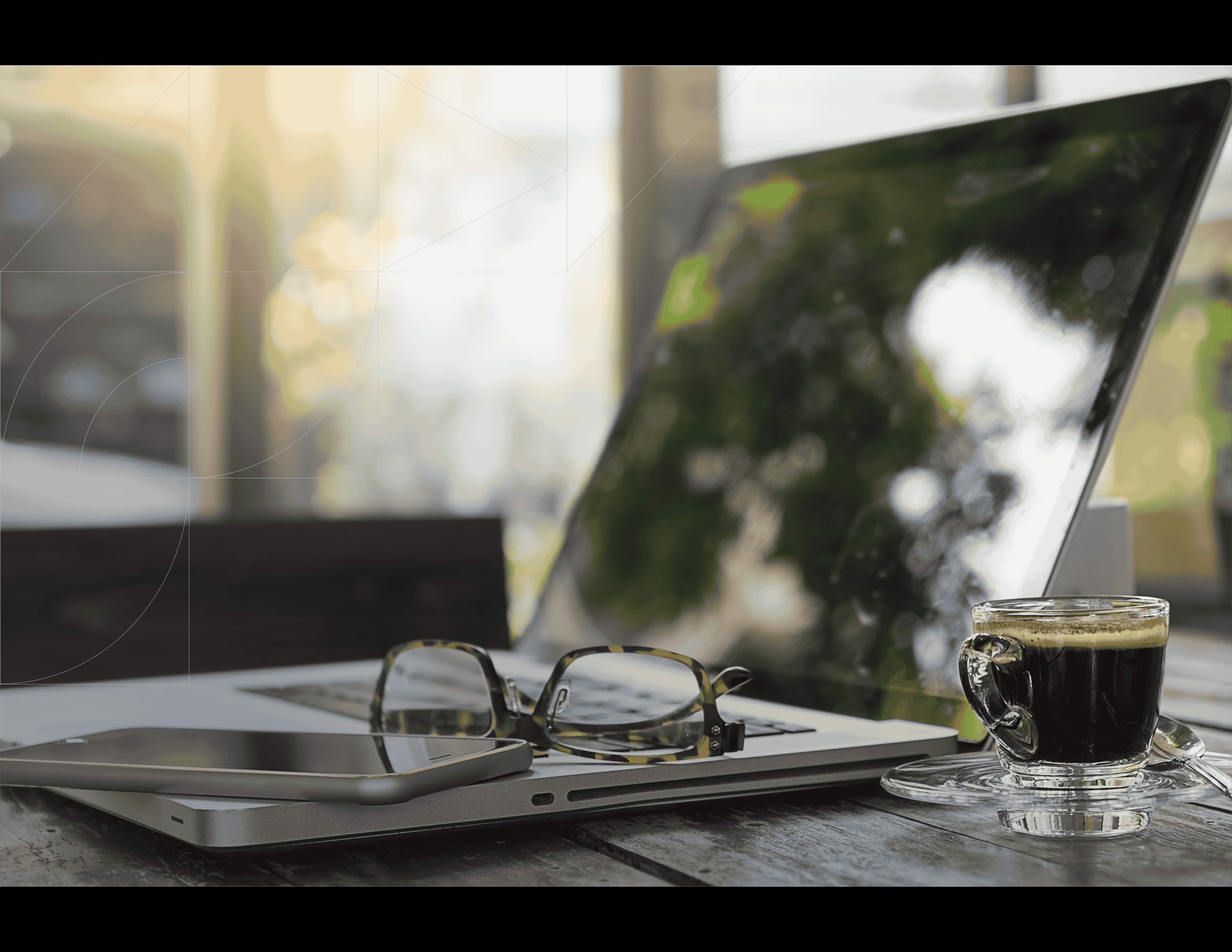 Laptop on a wooden table with glasses, a smartphone, and a cup of coffee in a bright workspace.