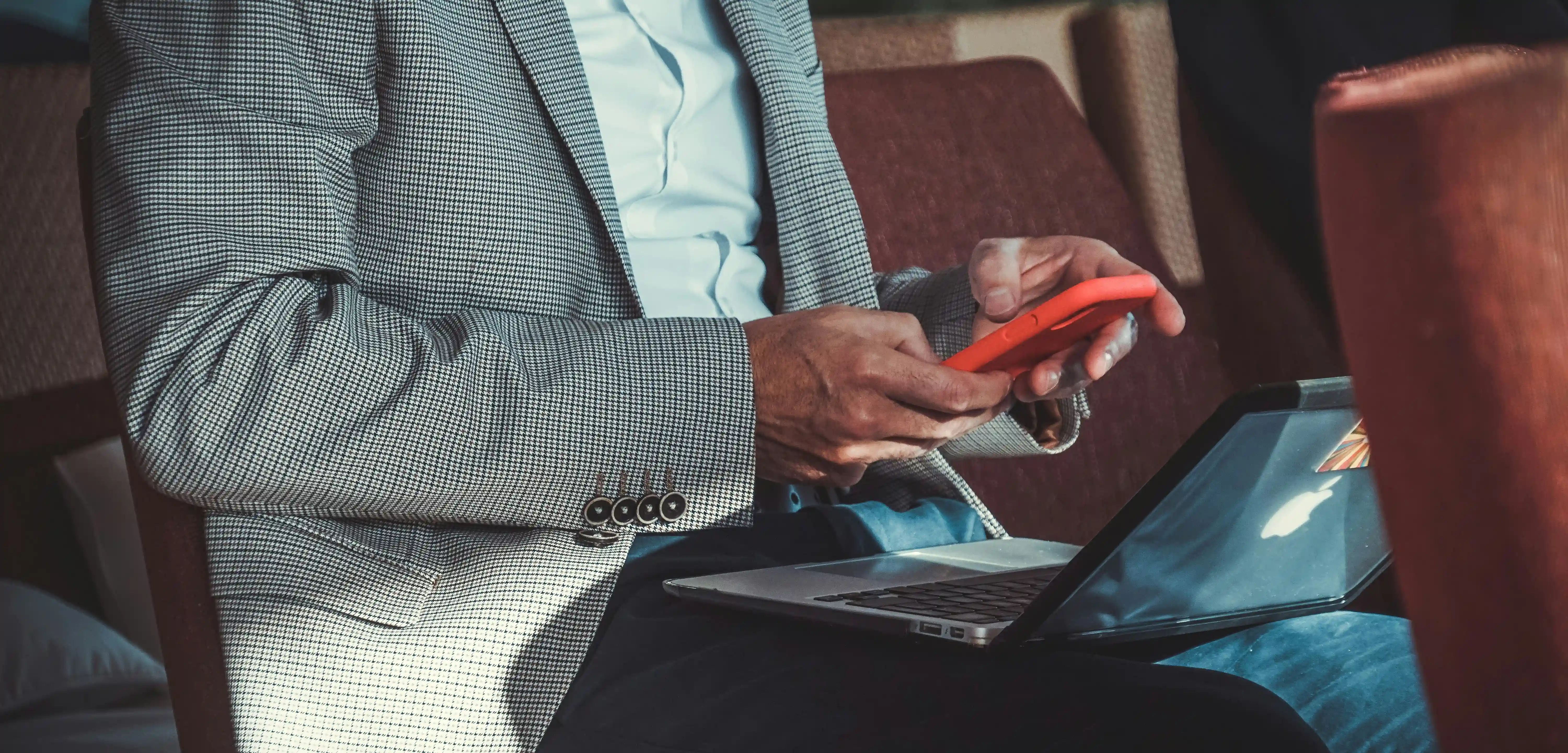 A person wearing a gray checkered blazer and blue pants is seated, using a red smartphone while a silver laptop rests open on their lap.