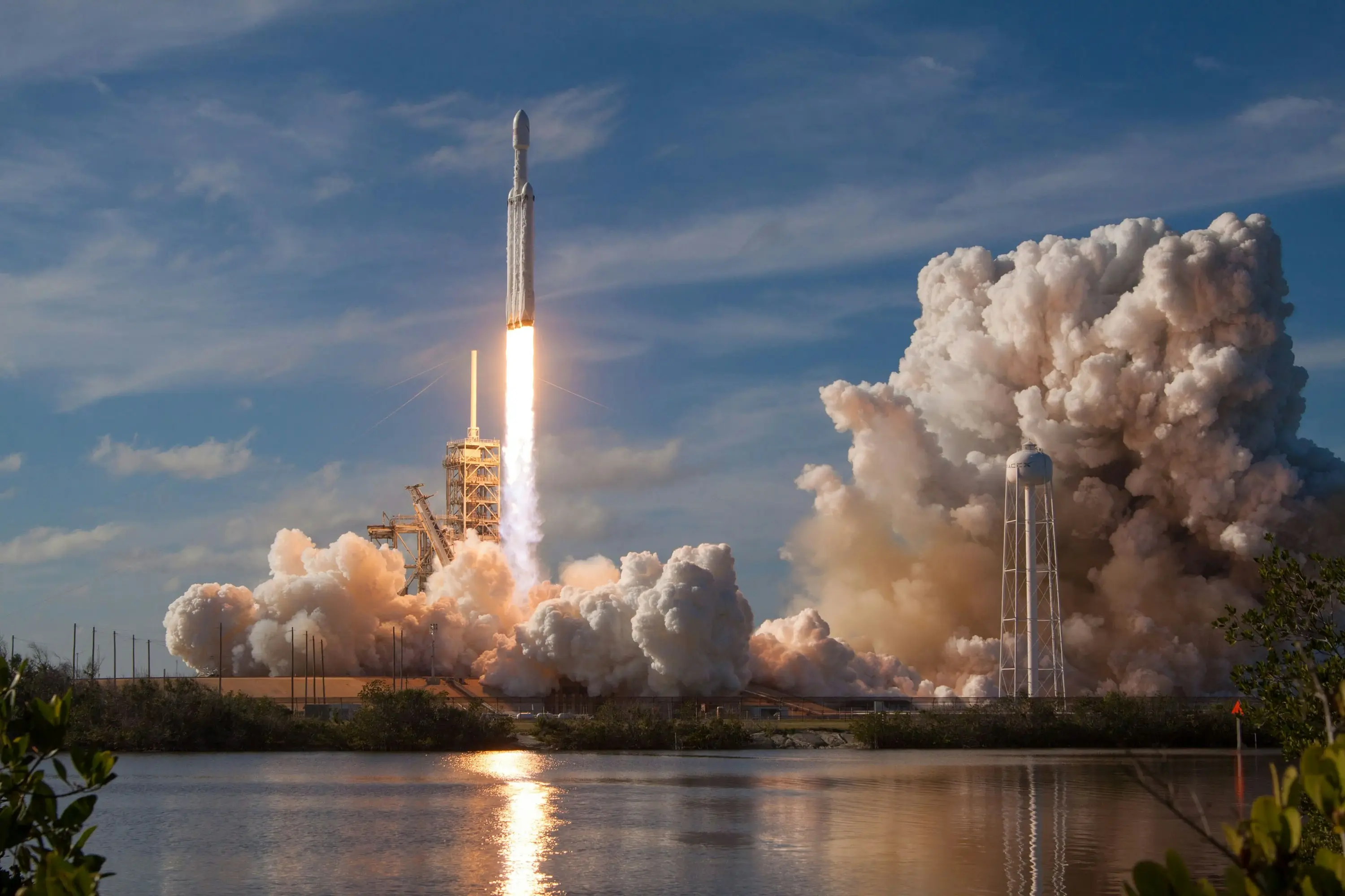 A rocket launches from a spaceport, blasting upward with a bright plume of fire and thick clouds of smoke billowing around the launch pad, reflected in a nearby body of water under a blue sky.
